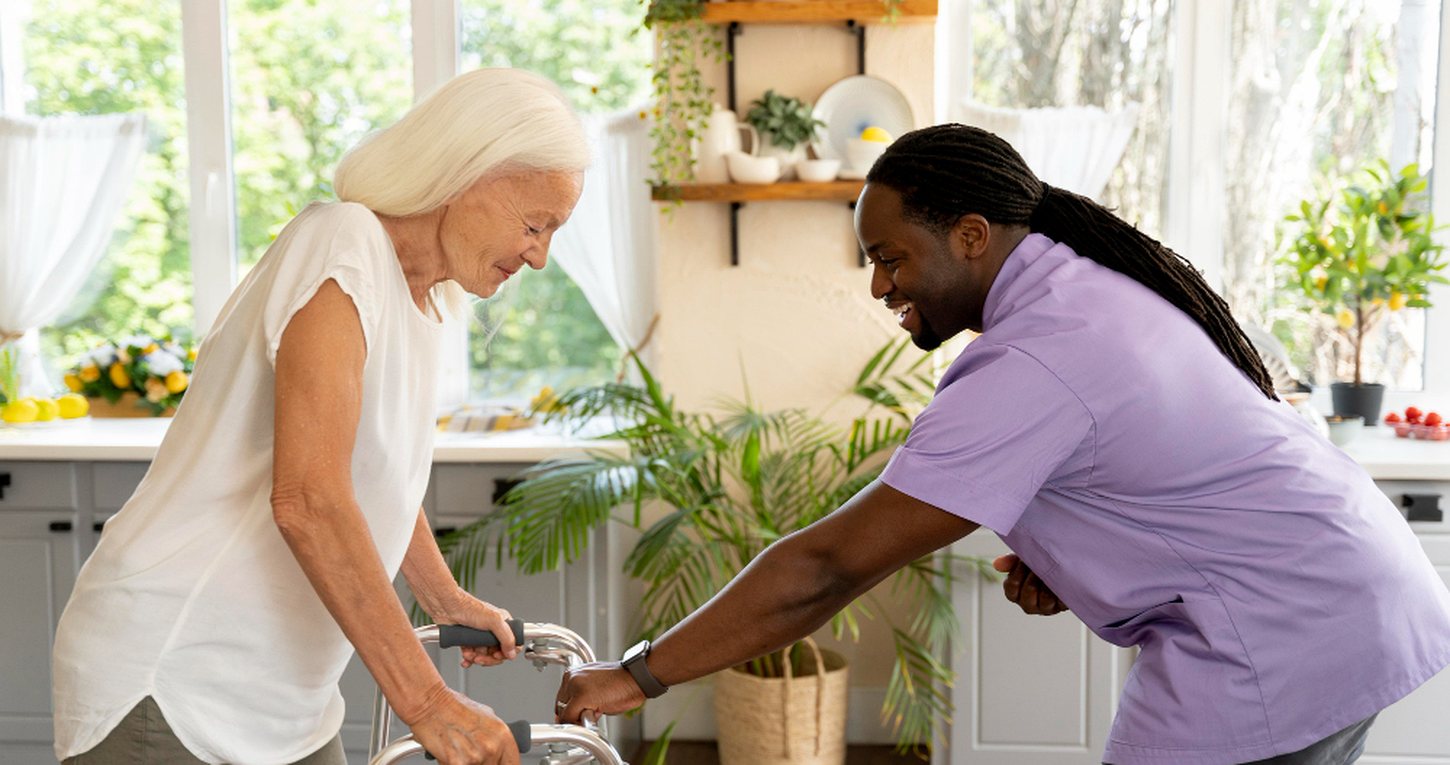 A caregiver steadying an older woman as she walks with a frame in her own kitchen